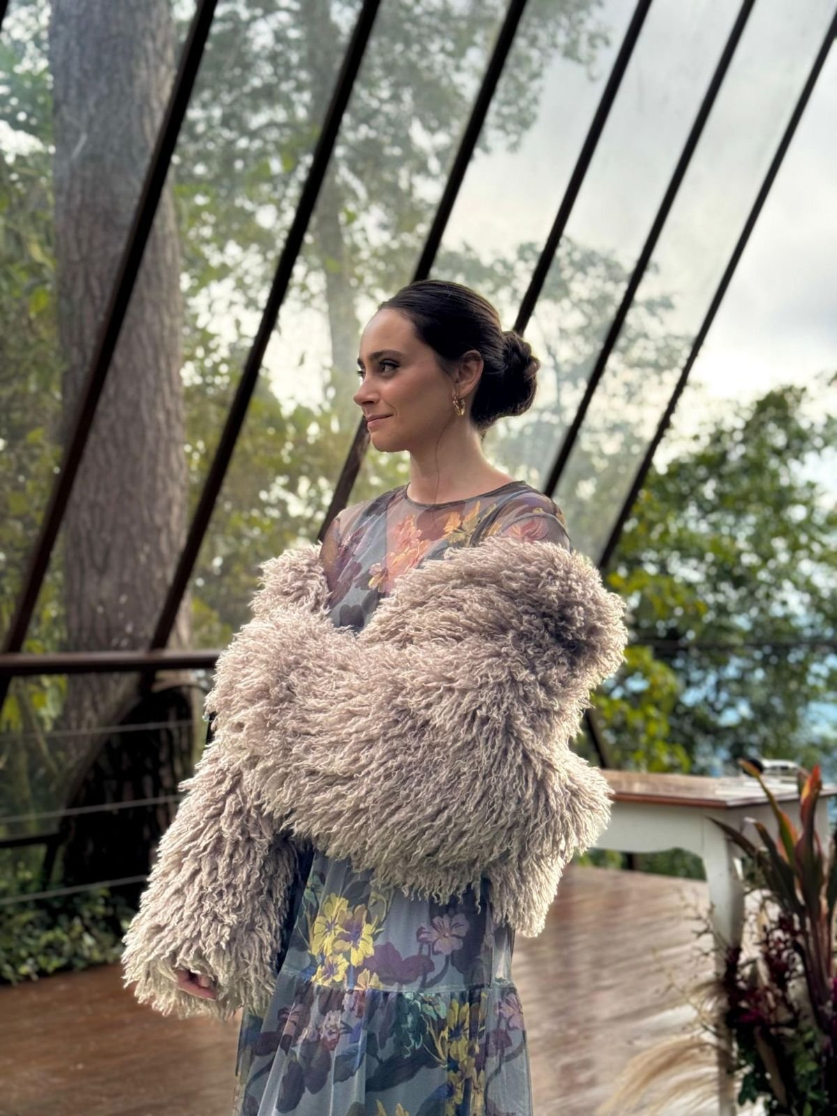 Mulher em vestido floral e casaco de lã sob uma estufa de vidro, usando brincos de argola dourados