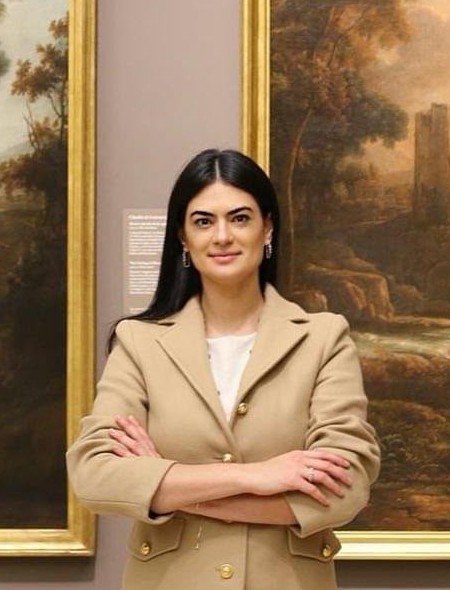 Jhess Alves de braços cruzados em um museu, entre duas pinturas de paisagem com molduras douradas